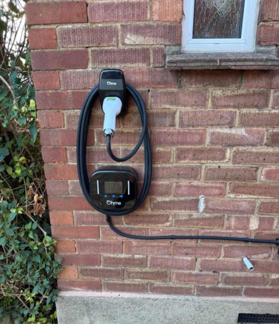 Ohme charge point on a red brick wall in Ruislip, charging lead coiled around the unit and the connector docked in a holster above.